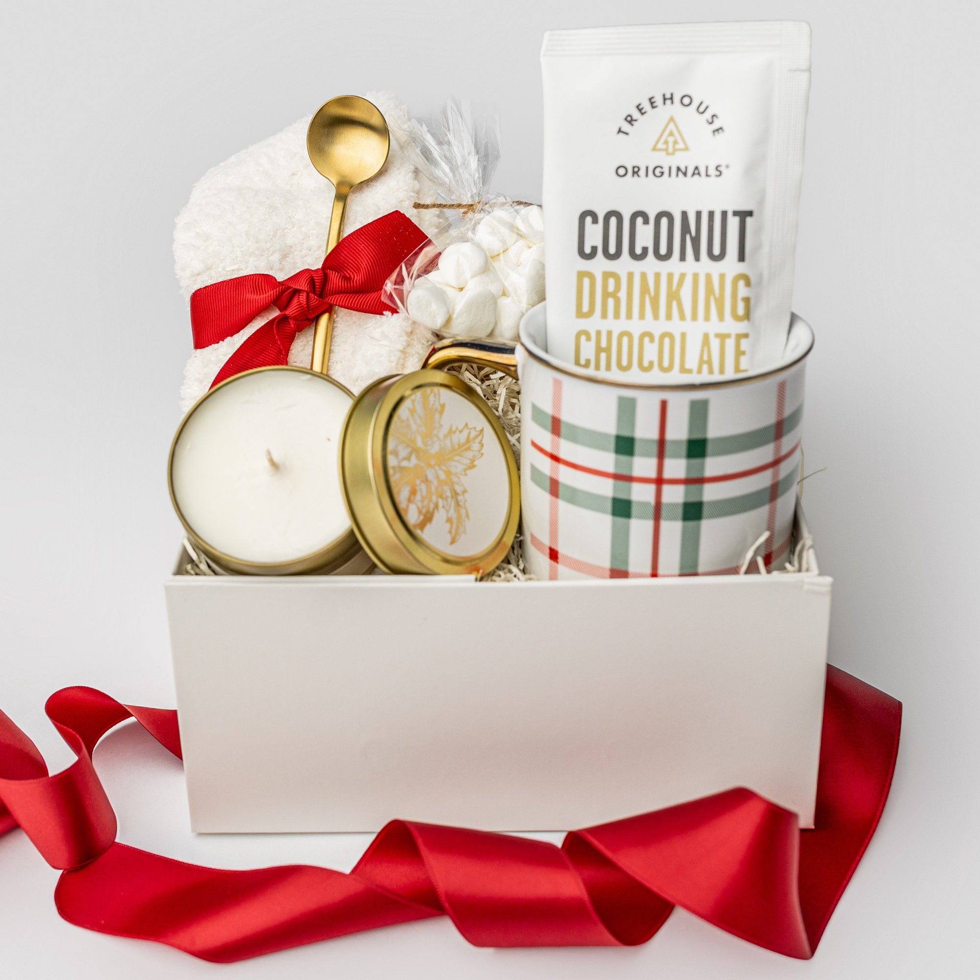 A white gift box filled with ivory paper shred, accented with a red ribbon around and under a pair of plush ivory cloud socks, on a white background. Inside the box are a 12 oz holiday plaid gold‑rimmed ceramic mug, a single‑serve packet of Treehouse Originals coconut hot cocoa, mini marshmallows, a 4 oz hand‑poured candle labeled “‘Tis the Season,” and a gold stirring spoon — a cozy holiday gift set.