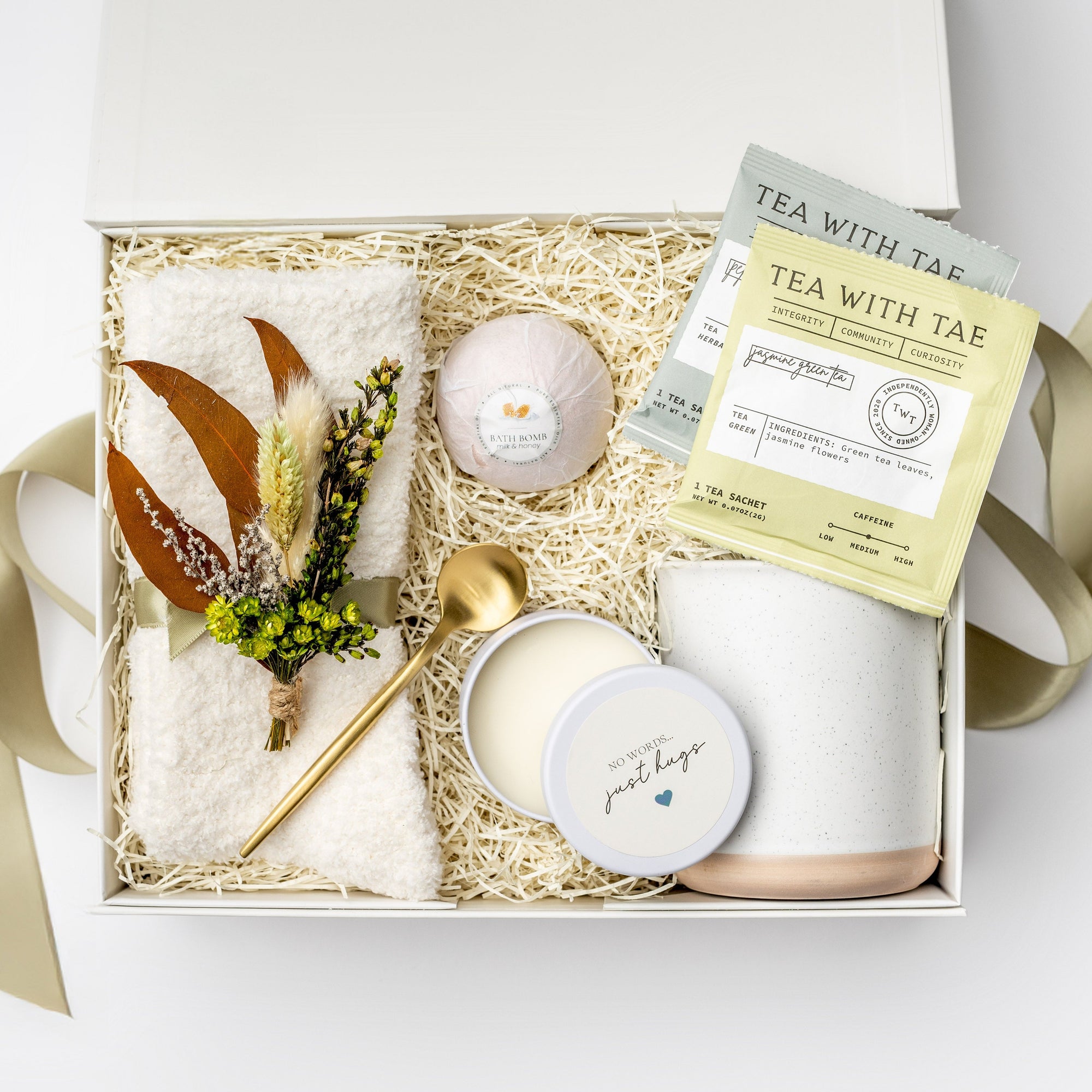 Warmest Wishes sympathy Gift Box presented in a white gift box filled with ivory shred and tied with a green ribbon, featuring a Milk &amp; Honey bath bomb, a 12 oz. speckled ceramic mug, two jasmine green and peppermint tea sachets, a gold stir spoon, ivory plush cloud socks, a 4 oz. hand-poured “No Words, Just Hugs” candle, and a handmade dried floral mini bouquet, all arranged on a white background.