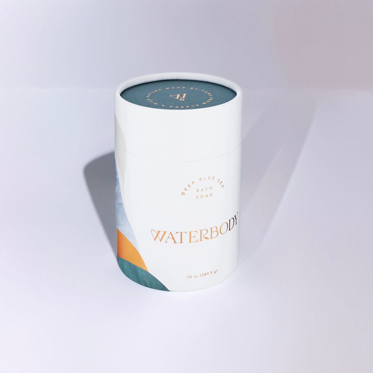 A white cylindrical bath bomb tin labeled WATERBODY Deep Blue Sea Bath Luxe, featuring a teal lid and abstract blue and orange geometric accents on the lower left, set against a clean white background with a soft shadow.
