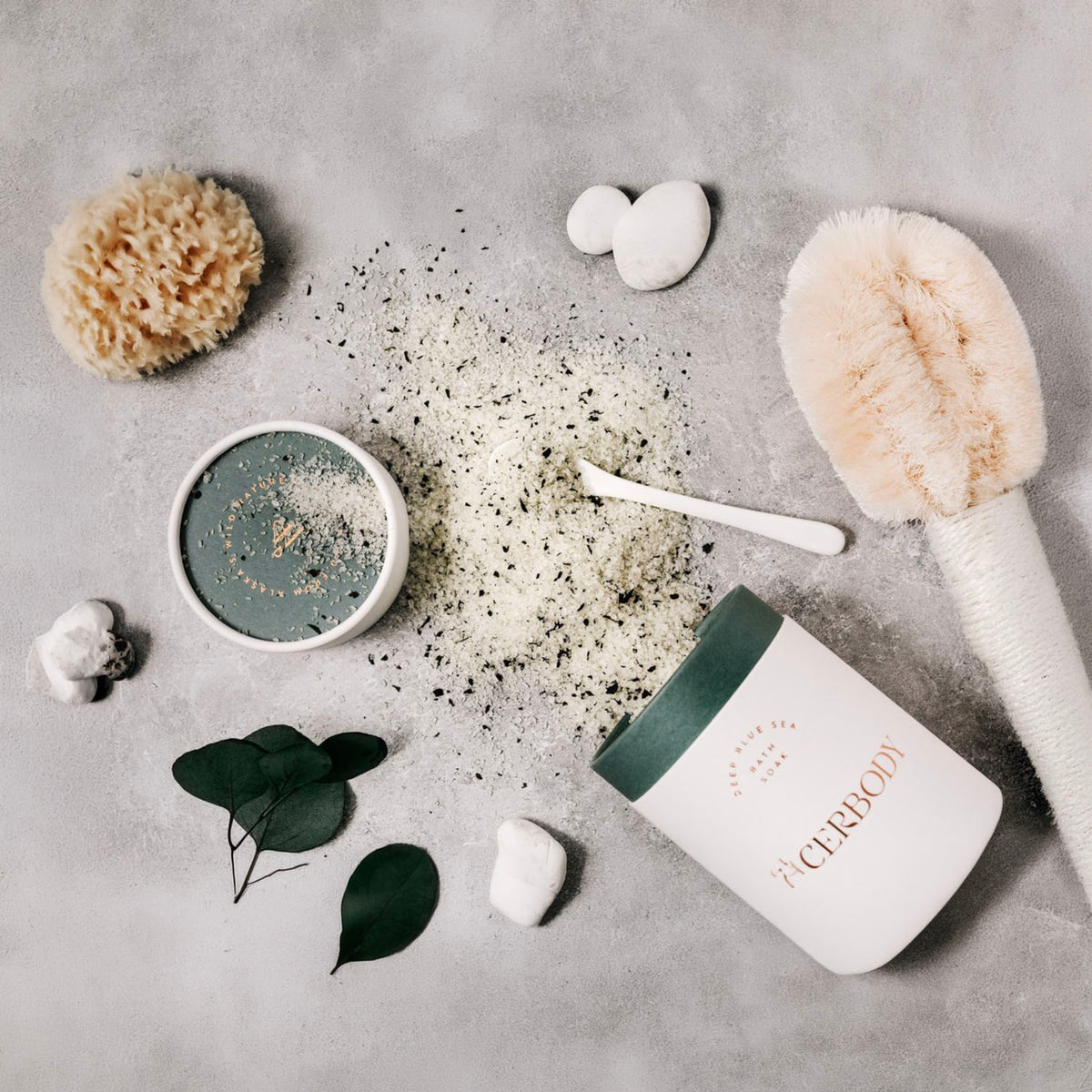 A flat-lay of a skincare product set on a textured gray background, featuring a white jar with a dark green lid labeled 'The Clerody' and 'The Clerydy' cream, alongside a small bowl of green scrub, loose powder, a sponge, a brush, and stones.