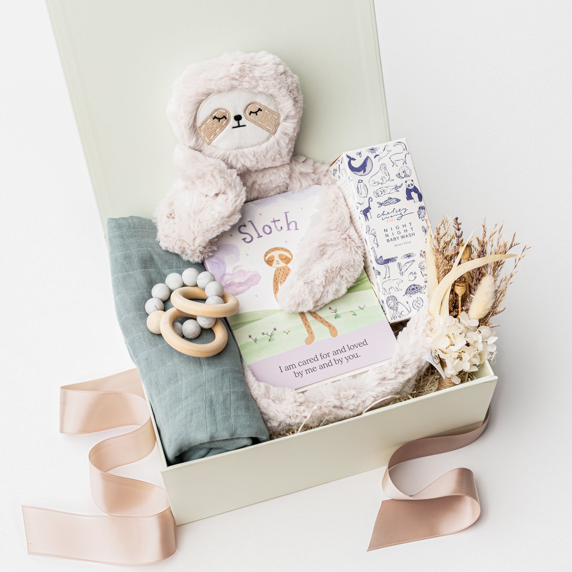 A top view of a baby gift set presented in a white gift box filled with ivory shred, featuring a Slumberkins Slumber Sloth, a muslin cotton swaddle blanket, Night Night baby wash, a silicone and wood baby teether, and a handcrafted dried floral mini bouquet, showing a tan ribbon, all arranged on a white background.