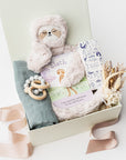 A top view of a baby gift set presented in a white gift box filled with ivory shred, featuring a Slumberkins Slumber Sloth, a muslin cotton swaddle blanket, Night Night baby wash, a silicone and wood baby teether, and a handcrafted dried floral mini bouquet, showing a tan ribbon, all arranged on a white background.