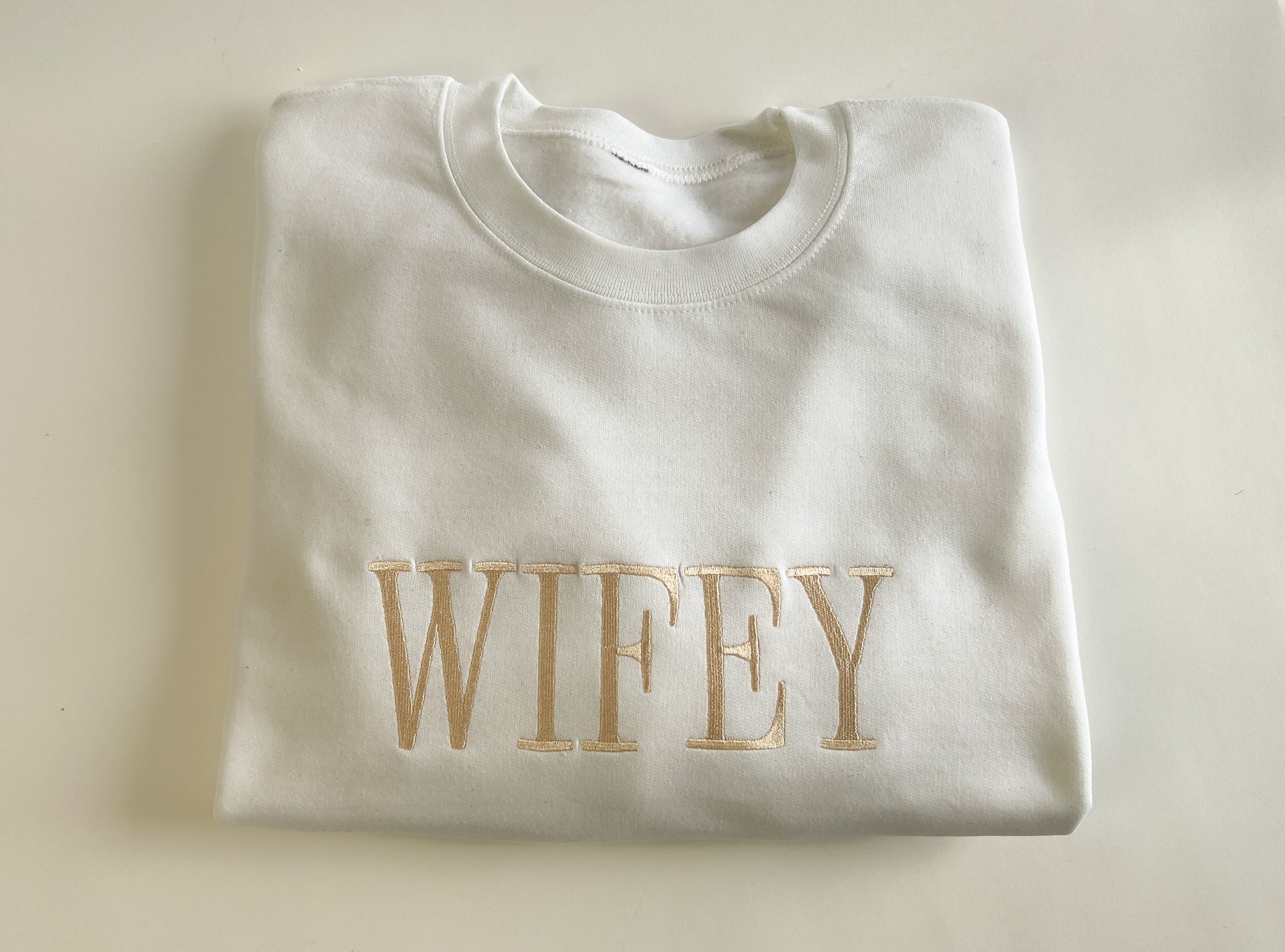 White crewneck sweatshirt featuring “wifey” embroidered in gold lettering, displayed on a white background.
