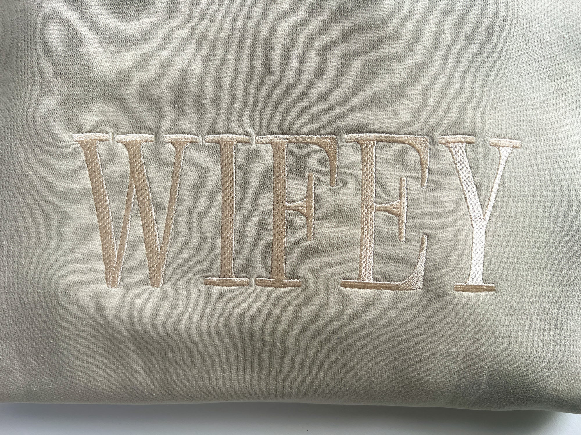 Close-up image of White crewneck sweatshirt featuring “wifey” embroidered in gold lettering, displayed on a white background.