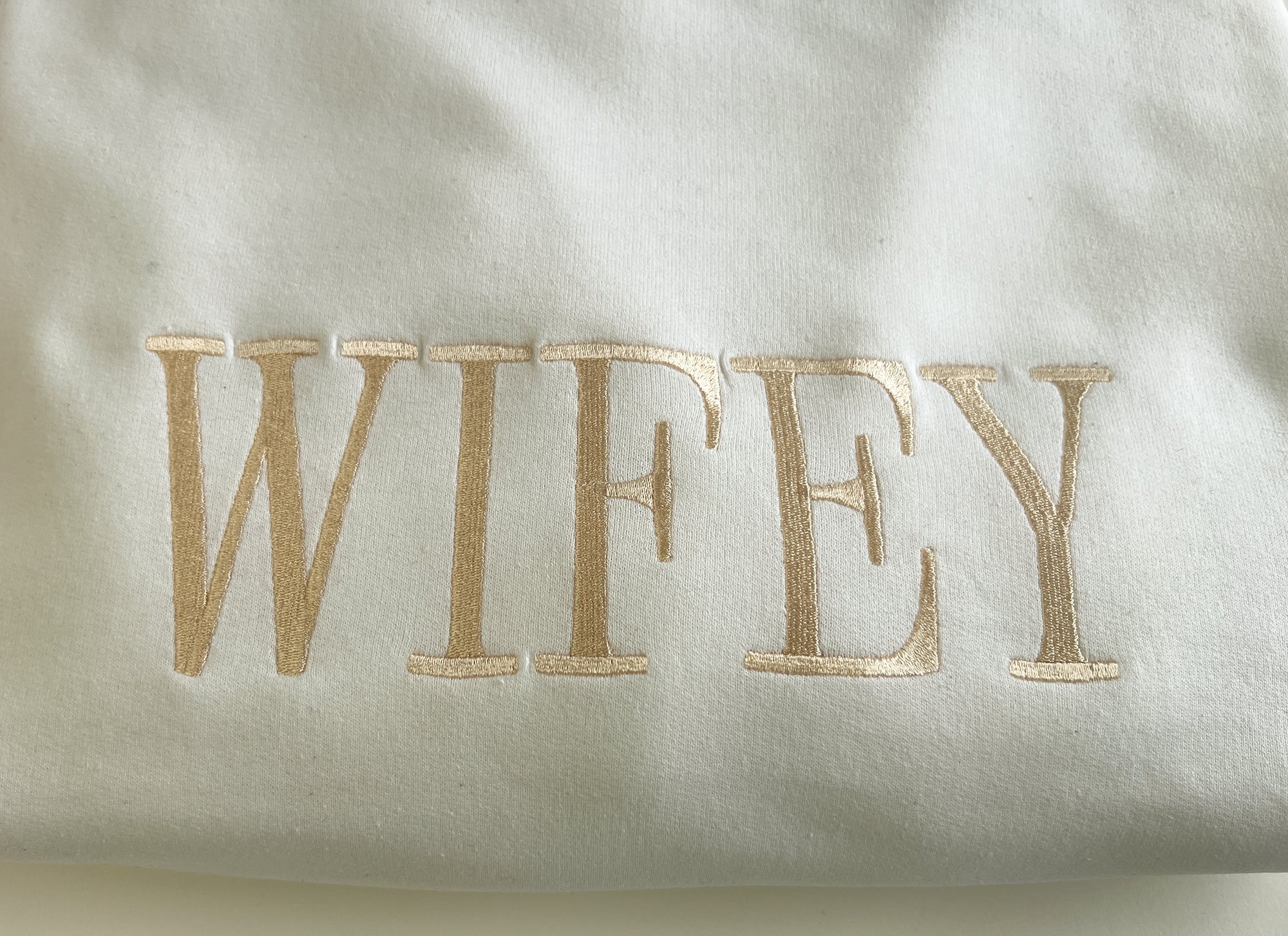 Close-up image of white crewneck sweatshirt featuring “wifey” embroidered in gold lettering, displayed on a white background.