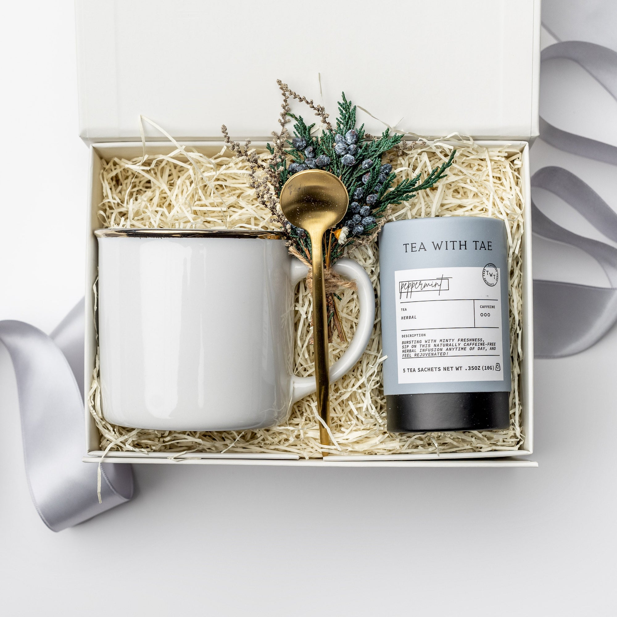 A white gift box with ivory paper shred and tied with a blue ribbon, set on a white background. Inside the box: a 12 oz ceramic gold-rimmed mug, a mini tube of artisan peppermint tea (5 sachets), a gold stirring spoon, and a handcrafted dried floral mini bouquet — a curated “Winter Tea” holiday gift box.
