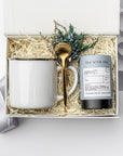 A white gift box with ivory paper shred and tied with a blue ribbon, set on a white background. Inside the box: a 12 oz ceramic gold-rimmed mug, a mini tube of artisan peppermint tea (5 sachets), a gold stirring spoon, and a handcrafted dried floral mini bouquet — a curated “Winter Tea” holiday gift box.
