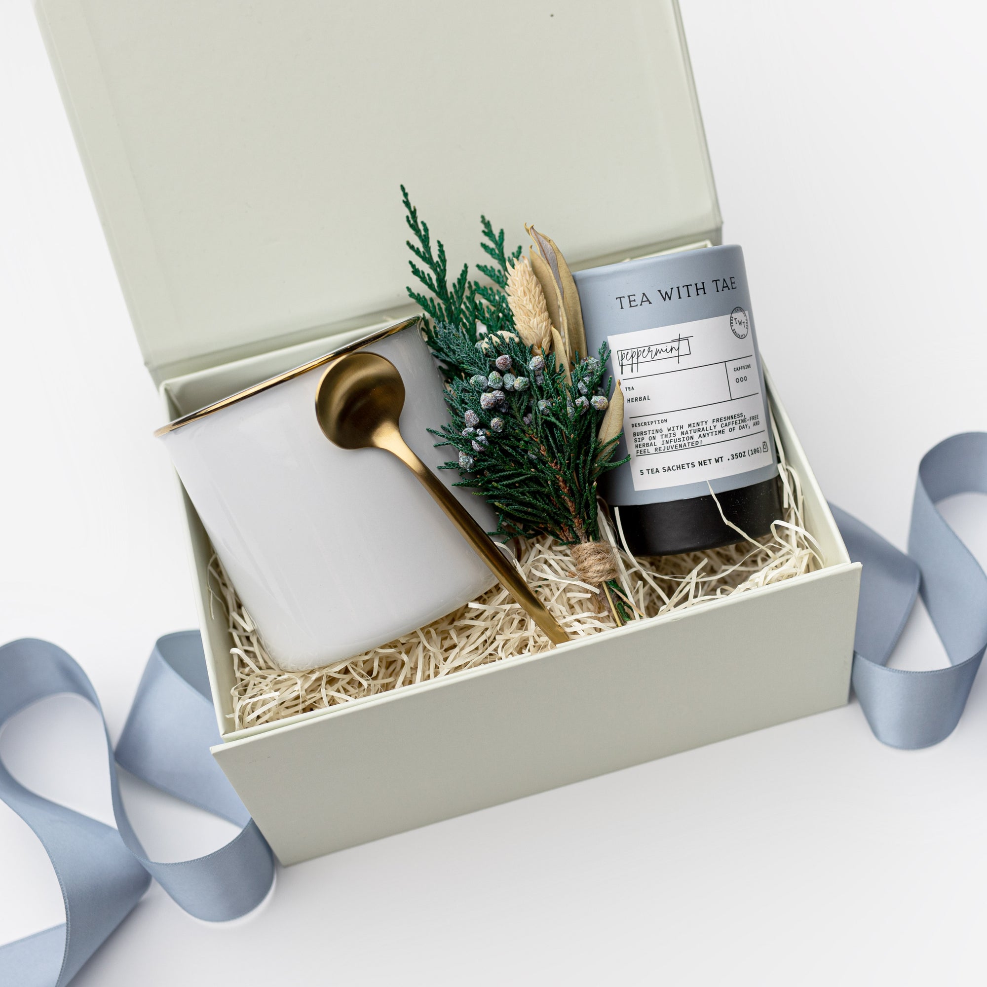 A white gift box with ivory paper shred and tied with a blue ribbon, set on a white background. Inside the box: a 12 oz ceramic gold-rimmed mug, a mini tube of artisan peppermint tea (5 sachets), a gold stirring spoon, and a handcrafted dried floral mini bouquet — a curated “Winter Tea” holiday gift box.
