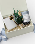 A white gift box with ivory paper shred and tied with a blue ribbon, set on a white background. Inside the box: a 12 oz ceramic gold-rimmed mug, a mini tube of artisan peppermint tea (5 sachets), a gold stirring spoon, and a handcrafted dried floral mini bouquet — a curated “Winter Tea” holiday gift box.
