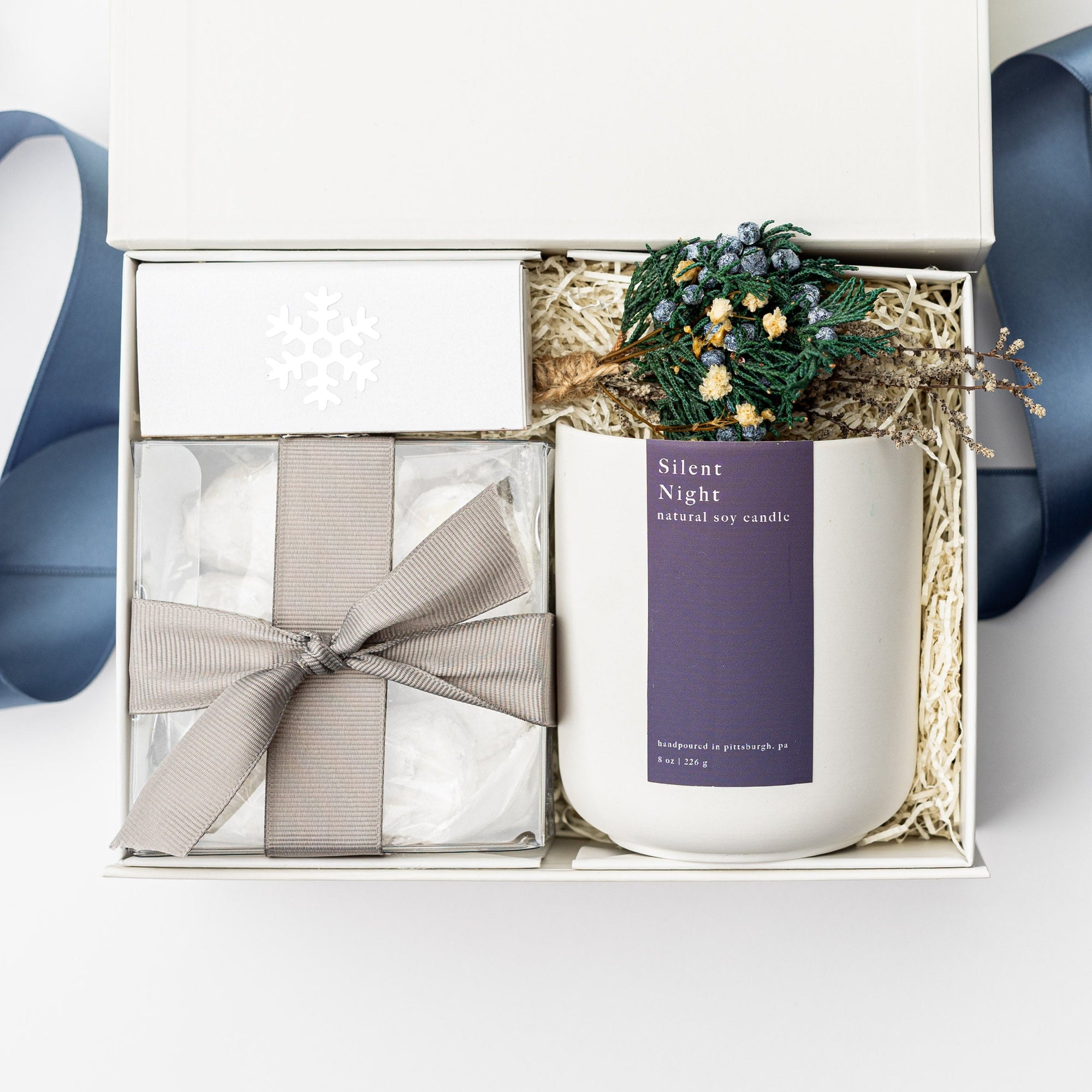 Above image of Winter Wonderland Gift Box featured with a white box containing a white ceramic “Silent Night” candle with a pblue label and white lettering, a handmade floral mini bundle with blue, yellow, green, and beige flowers wrapped in a twig, a box of tea cakes in a transparent square container tied with a silver ribbon, and a snowflake silver foil candle matchbox. The box is filled with ivory shred in the back and finished with a blue ribbon in the back, displayed on a white background.