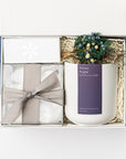 Above image of Winter Wonderland Gift Box featured with a white box containing a white ceramic “Silent Night” candle with a pblue label and white lettering, a handmade floral mini bundle with blue, yellow, green, and beige flowers wrapped in a twig, a box of tea cakes in a transparent square container tied with a silver ribbon, and a snowflake silver foil candle matchbox. The box is filled with ivory shred in the back and finished with a blue ribbon in the back, displayed on a white background.