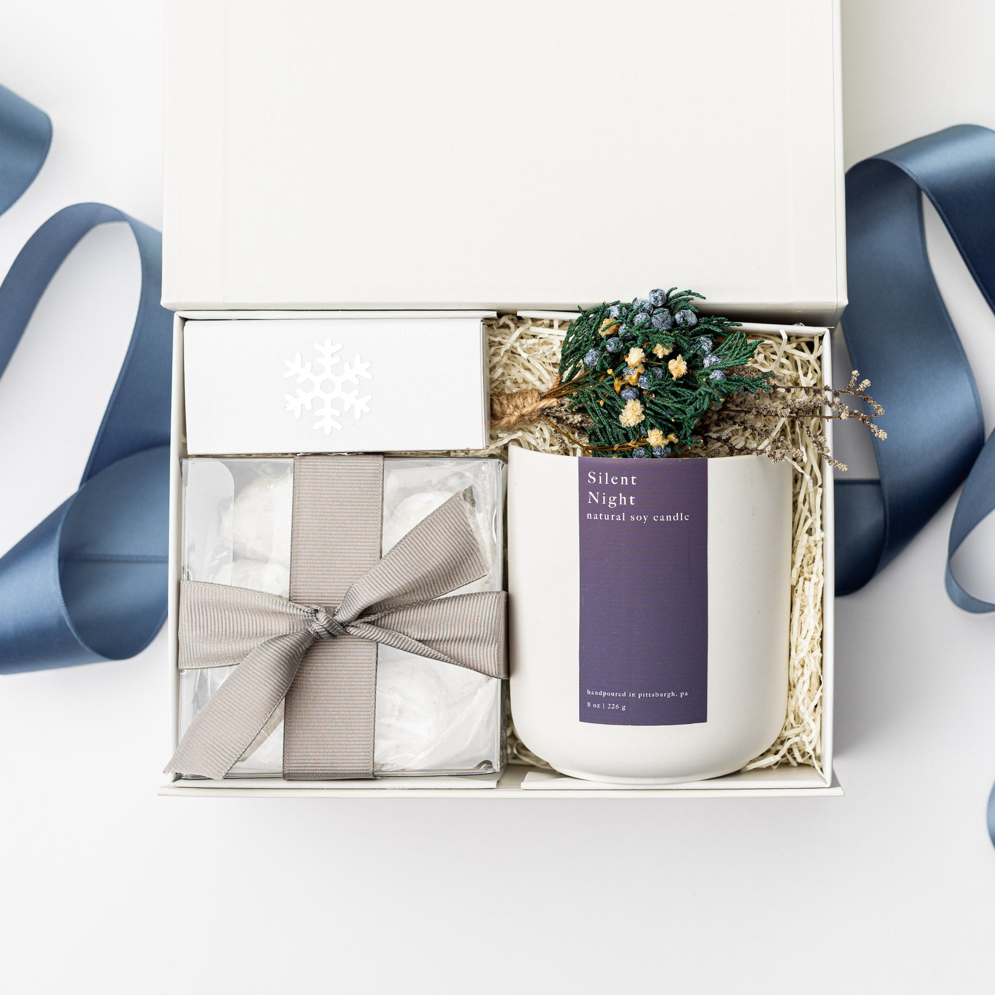 Above image of Winter Wonderland Gift Box featured with a white box containing a white ceramic “Silent Night” candle with a purple label and white lettering, a handmade floral mini bundle with blue, yellow, green, and beige flowers wrapped in a twig, a box of tea cakes in a transparent square container tied with a silver ribbon, and a snowflake silver foil candle matchbox. The box is filled with ivory shred in the back and finished with a blue ribbon in the back, displayed on a white background.