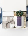 Above image of Winter Wonderland Gift Box featured with a white box containing a white ceramic “Silent Night” candle with a purple label and white lettering, a handmade floral mini bundle with blue, yellow, green, and beige flowers wrapped in a twig, a box of tea cakes in a transparent square container tied with a silver ribbon, and a snowflake silver foil candle matchbox. The box is filled with ivory shred in the back and finished with a blue ribbon in the back, displayed on a white background.