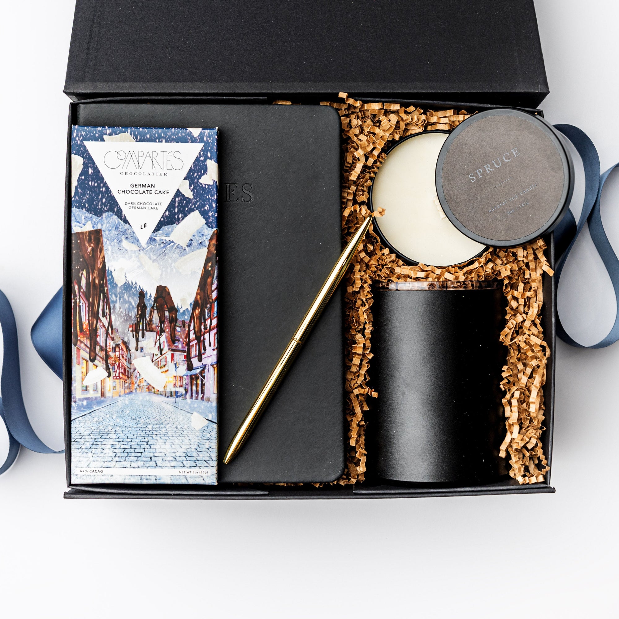 A black gift box filled with brown paper shred and tied with a blue ribbon on a white background. Inside the box are a black softcover notebook, a gold minimalist pen, a Compartes German Chocolate Cake bar, an 8 oz hand-poured soy candle in the scent “Spruce,” and a 12 oz vacuum-insulated travel mug — a “Winter Work From Home” gift box designed for productivity and comfort.