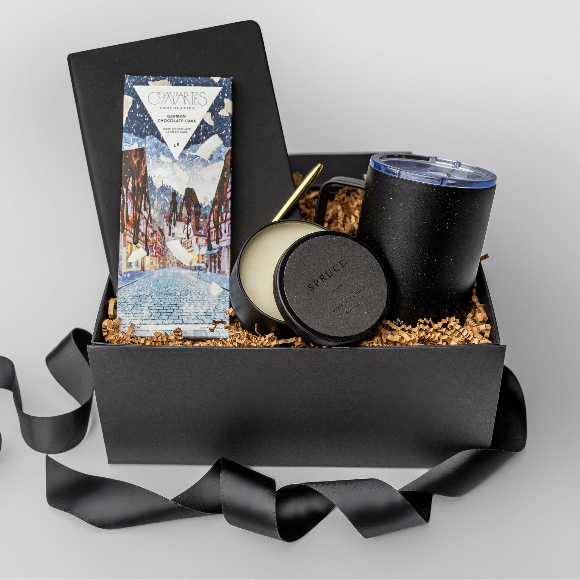 A black gift box filled with brown paper shred and tied with a blue ribbon on a white background. Inside the box are a black softcover notebook, a gold minimalist pen, a Compartes German Chocolate Cake bar, an 8 oz hand-poured soy candle in the scent “Spruce,” and a 12 oz vacuum-insulated travel mug — a “Winter Work From Home” gift box designed for productivity and comfort.