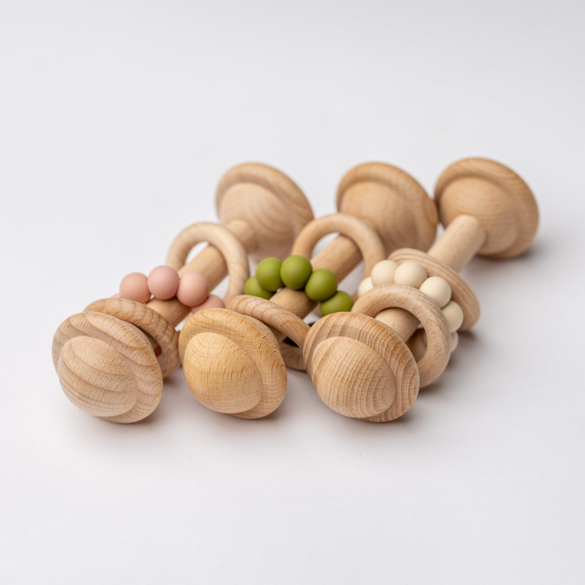 Eco-friendly wooden baby rattle with a smooth maple wood handle and simple natural design for infant play, three shown, each with a different color of silicone beads: pink, green, and beige.