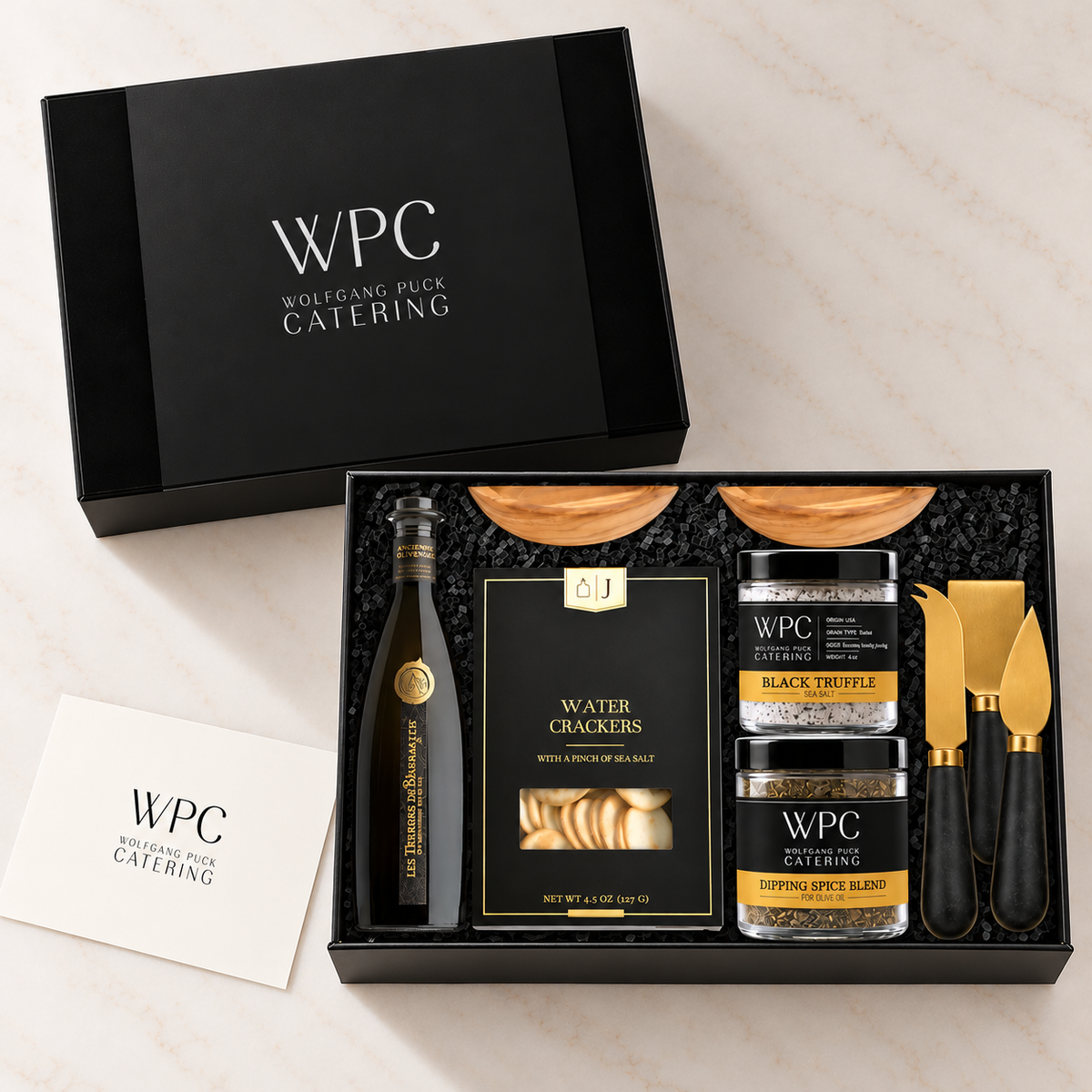 WPC Wolfgang Puck Catering gift set displayed in an open black presentation box on a light marble surface, with the matching closed box shown above. The set includes a bottle of oil, water crackers, black truffle and peppercorn mixes, wooden serving boards, and gold-handled serving utensils.