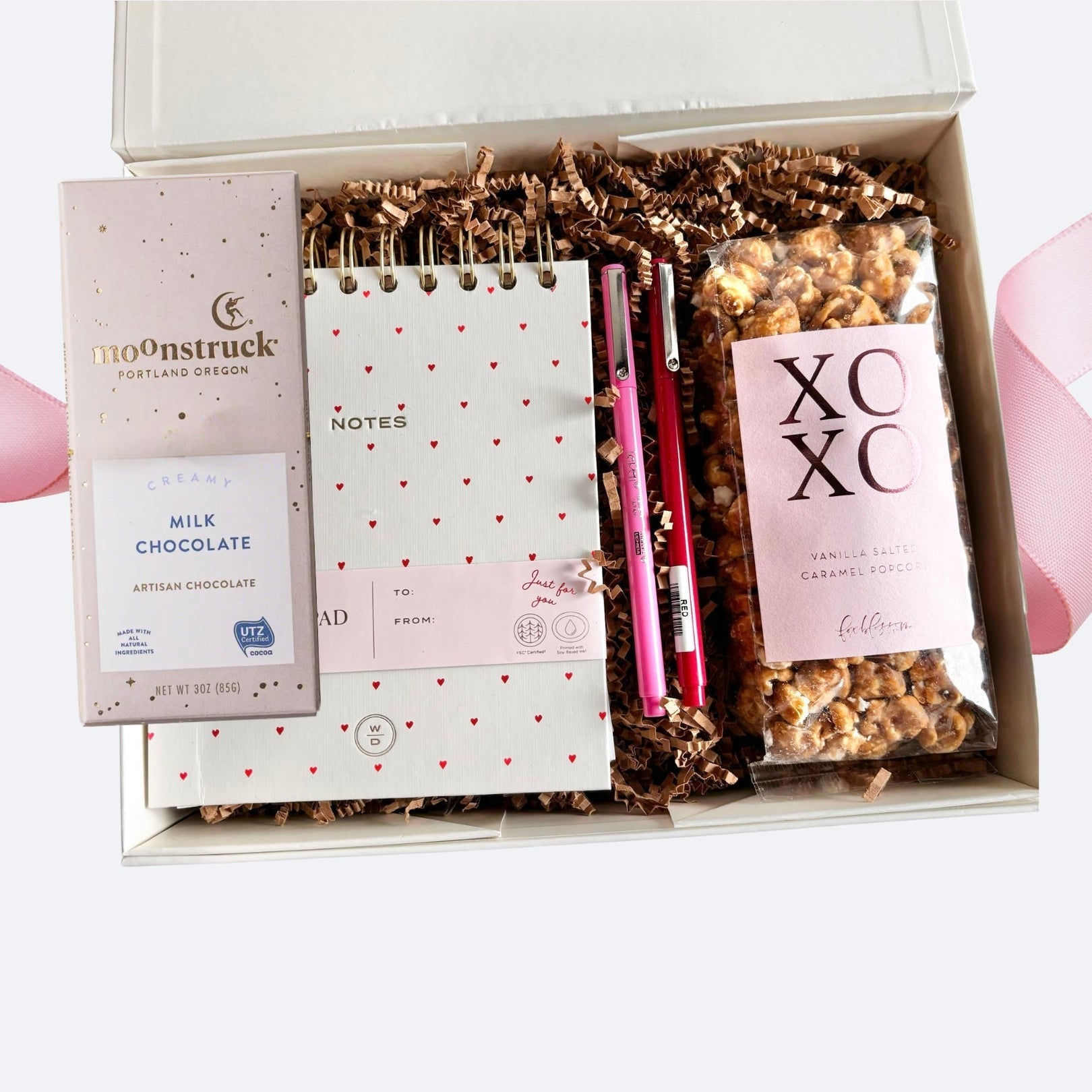 Open white gift box with assorted items including a bar of Moonstruck creamy milk chocolate in pink wrapping, a small spiral white notebook with a red hearts design, one pink pen, one red pen, and a bag of vanilla salted caramel popcorn labeled ‘XOXO,’ all arranged on brown crinkle paper.
