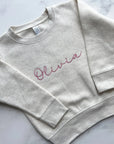 Youth custom embroidered sweatshirt cream with pink “Olivia” script on a marble background.