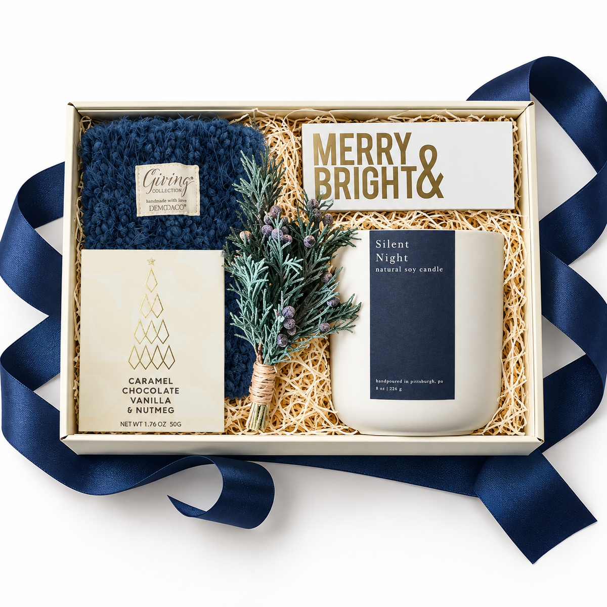 A curated gift box set on a white background, featuring a navy blue knit blanket, a bar of Caramel Chocolate, a 'Merry &amp; Bright' sign, and a 'Silent Night' soy candle nestled in shredded paper with a small pine sprig and navy ribbon.