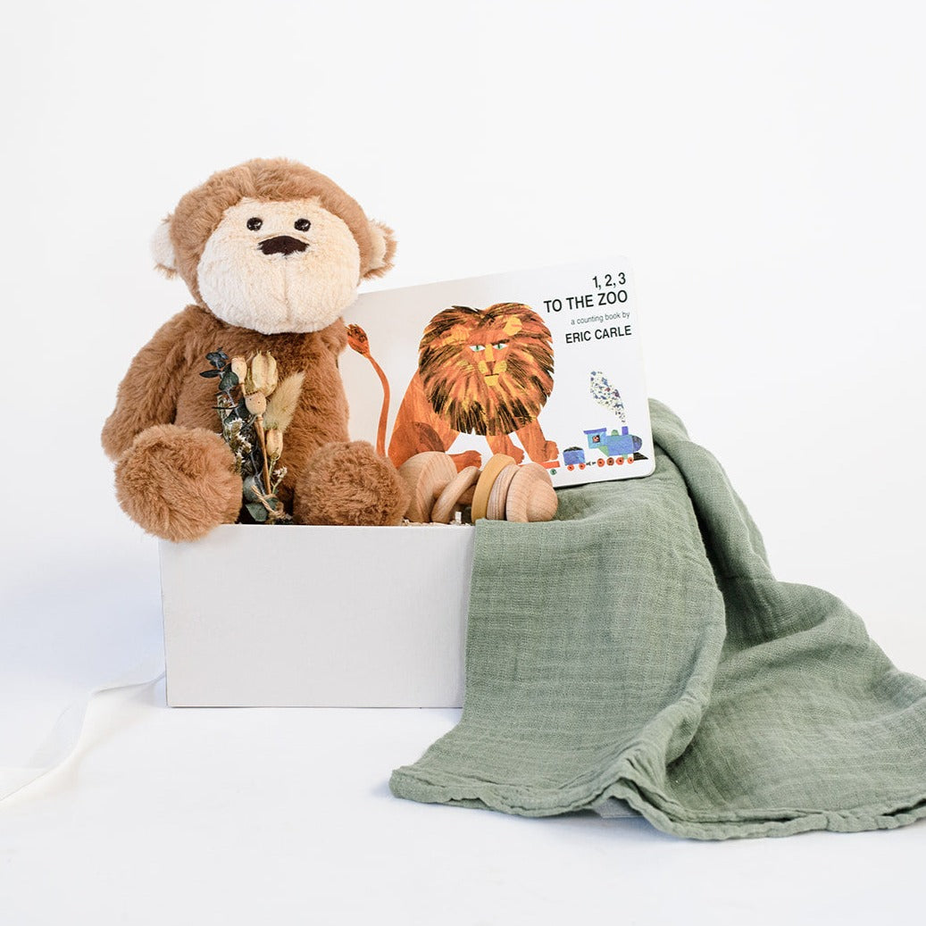Light grey gifting box with Manhattan Toy Co brown Mocha monkey plush sitting with dried floral bundle, Eric Carle's board book "1, 2, 3 To The Zoo," showing a lion riding a small train, light wood teething rattle with a yellow silicon ring, and sage green muslin swaddle.
