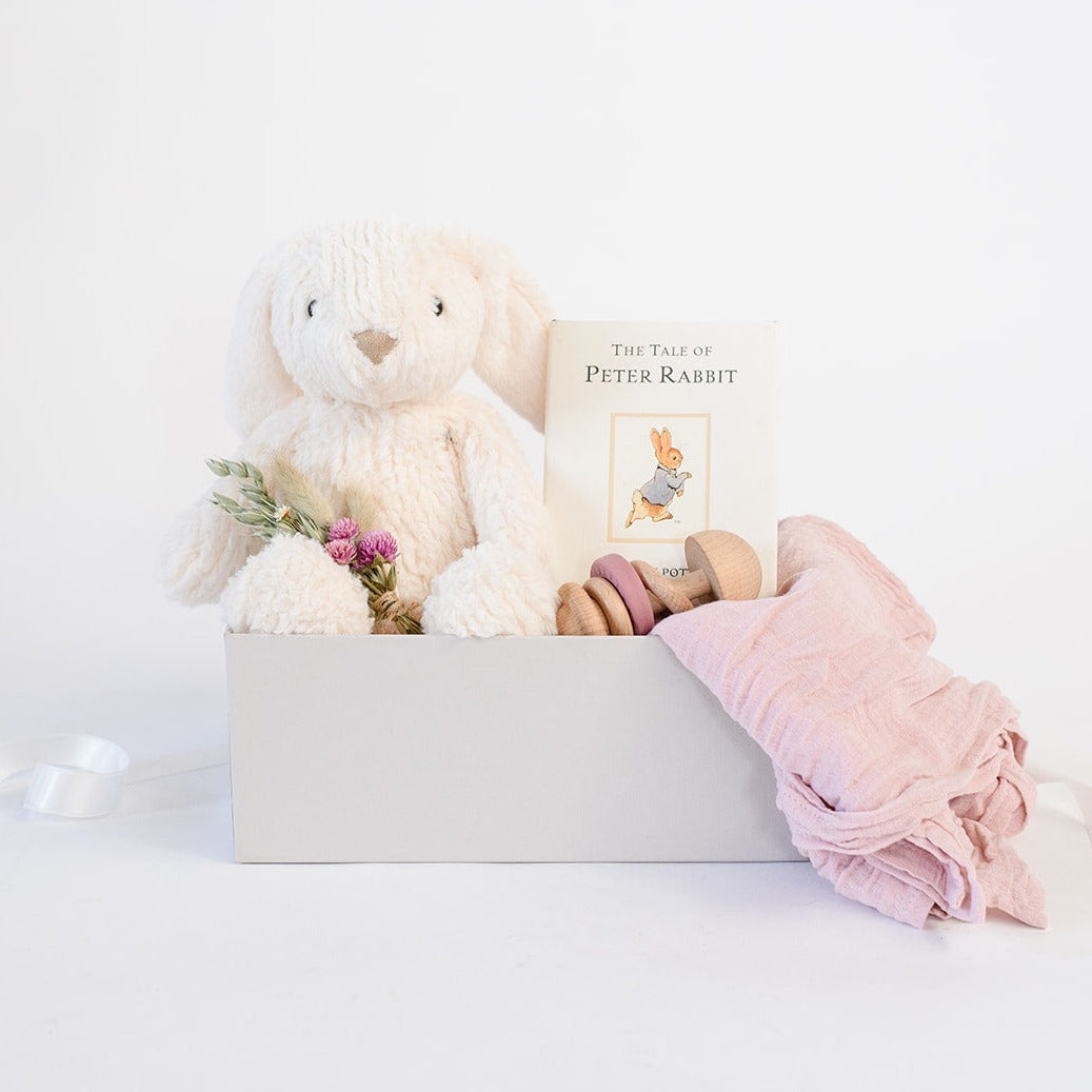 White Lulu Rabbit plush from Manhattan Toy Company, pink dried floral bundle, Peter Rabbit hardcover book, wooden teething rattle with pink silicone, light pink muslin swaddle. All in grey gifting box 