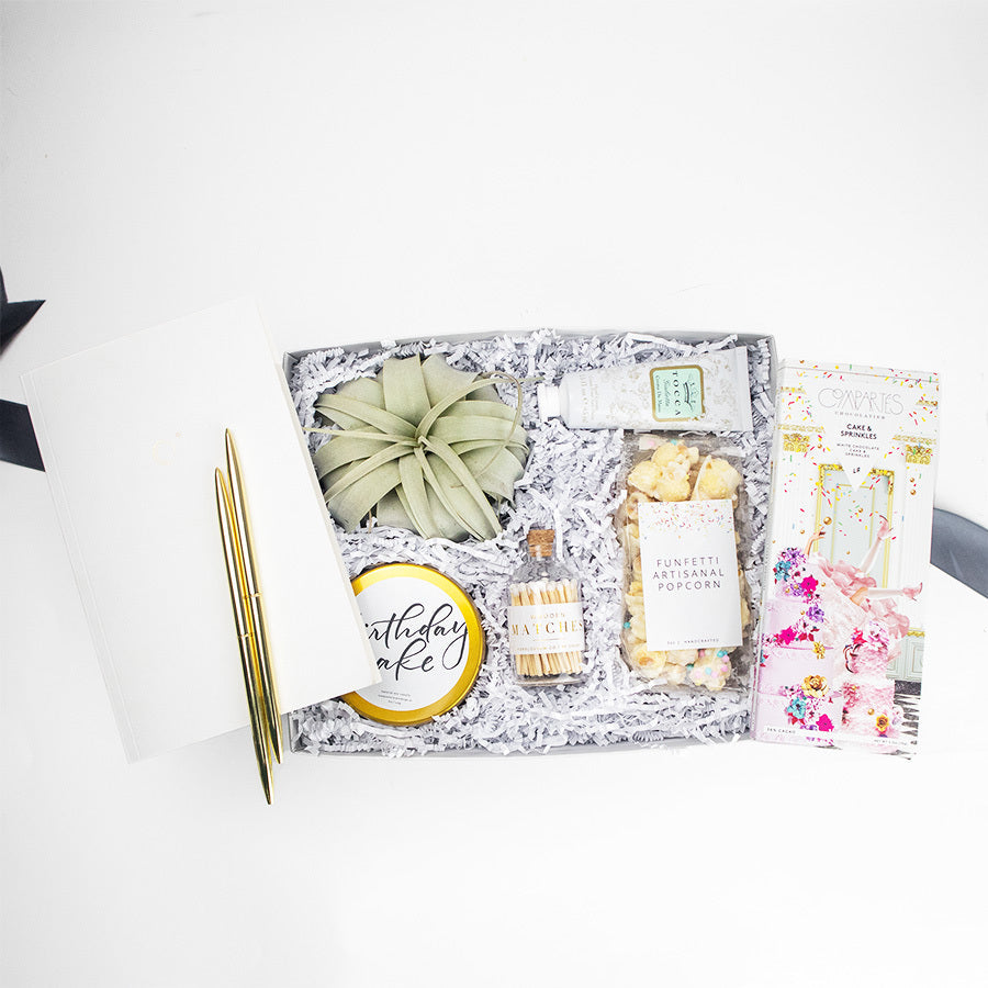 Light grey box containing a large air plant, bottle of white tipped matches, two gold pens, a compartes "cake & sprinkles" chocolate bar, birthday cake candle in gold tin, white journal, mini tube of Tocca Giulietta hand cream and clear bag of funfetti artisanal popcorn.