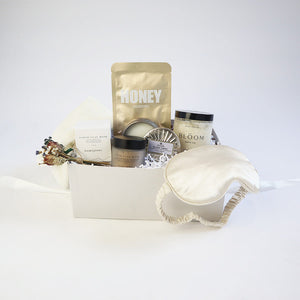 Light grey box containing ivory chenille cozy socks, earth clay mask in opaque frosted jar with black lid and white box, "honey nourishing" sheet mask in golden foil packet, antiqued silver blond tabac candle next to matching lid, clear jar of "Bloom bath co" grapefruit scrub, champagne satin sleep mask. 