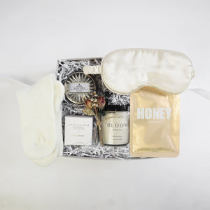 Ivory chenille cozy socks, earth clay mask in opaque frosted jar with black lid and white box, "honey nourishing" sheet mask in golden foil packet, antiqued silver blond tabac candle next to matching lid, clear jar of "Bloom bath co" grapefruit scrub, champagne satin sleep mask arranged in a light grey box with white paper shred.
