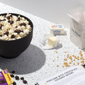Black bowl filled with popcorn and raisinets  next to sticks of butter, sea salt, popcorn, a box of raisinets and a book open to a page that read "popcorn with chocolate covered raisins.