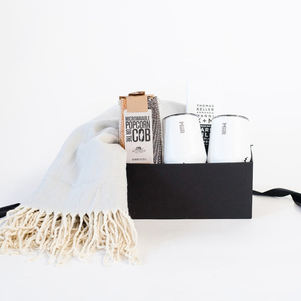 Black box containing a grey herringbone throw blanket, two ears of microwavable corn on the cob, two white insulated stemless tumblers, and dark milk chocolate bar. 