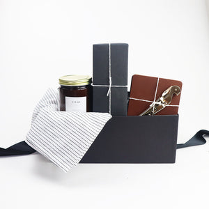 Black box containing a white linen tea towel with black pinstripes, Chai candle in an amber jar with gold lid, a box of rustic baker pecan shortbread cookies wrapped in black paper, set of four leather coasters and a gold plated corkscrew.