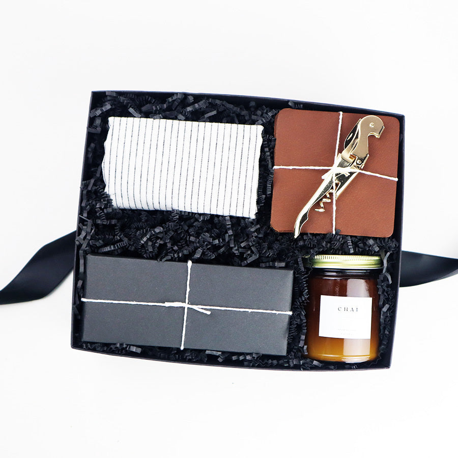 Black box containing a white linen tea towel with black pinstripes, Chai candle in an amber jar with gold lid, a box of rustic baker pecan shortbread cookies wrapped in black paper, set of four leather coasters and a gold plated corkscrew.