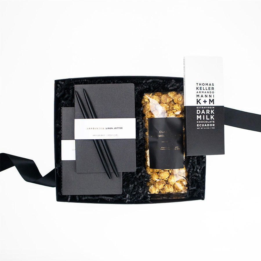 A black gifting box with a K+M Extravirgin Dark Milk Chocolate Ecuador bar, Large vanilla caramel popcorn with a black and gold label, two black pens, and two Appointed dark grey linen jotter journals.