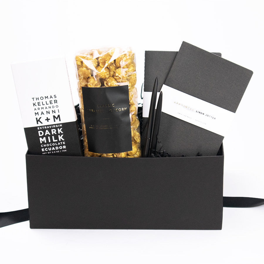 A black gifting box with a K+M Extravirgin Dark Milk Chocolate Ecuador bar, Large vanilla caramel popcorn with a black and gold label, two black pens, and two Appointed dark grey linen jotter journals.