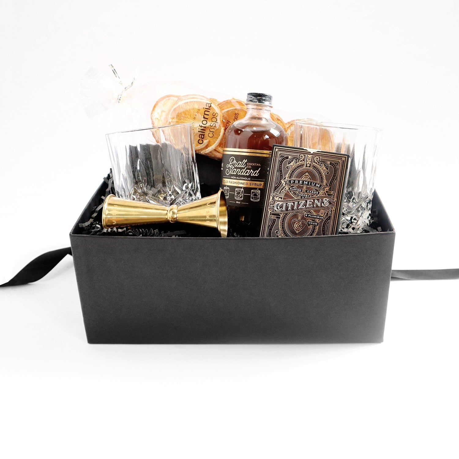 Black box with a set of two crystal tumblers, a double sided golden jigger, bottle of old fashioned syrup with black and gold label, a pack of playing cards  with elaborate gold and bronze foil design, pack of dried oranges.