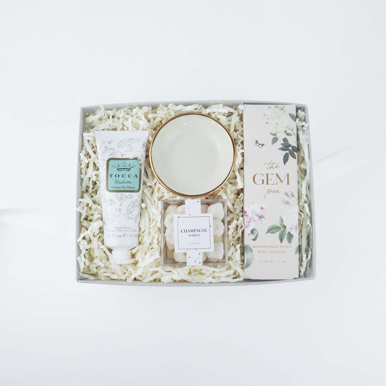 Light grey box containing a small, white ceramic ring dish with gold rim, floral filigree tube of Tocca Giullietta hand cream, white "Gem Pen" with floral design and matching box, clear plastic "champagne bubbles" cube containing gummy drops covered with white non-pareils. 