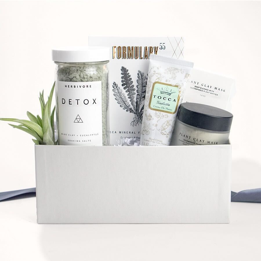 A light grey gift box with Herbivore Detox soaking salts, Formulary 55 sea mineral foot soak, Tocca guilietta hand cream, a Nash Jones plant clay mask, and a small airplant. 