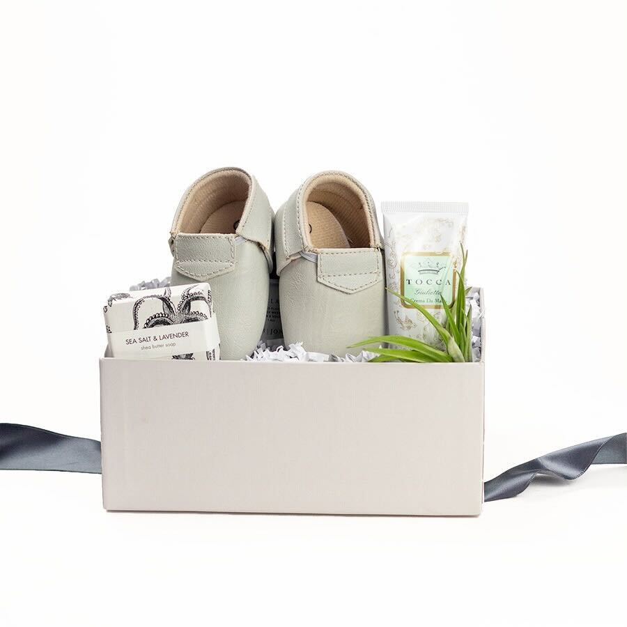 Light grey gift box with Sweet n' Swag modern mox baby shoes, Tocca Giulietta hand cream, a small air plant, and Formulary 55 Sea Salt & Lavender shea butter soap.