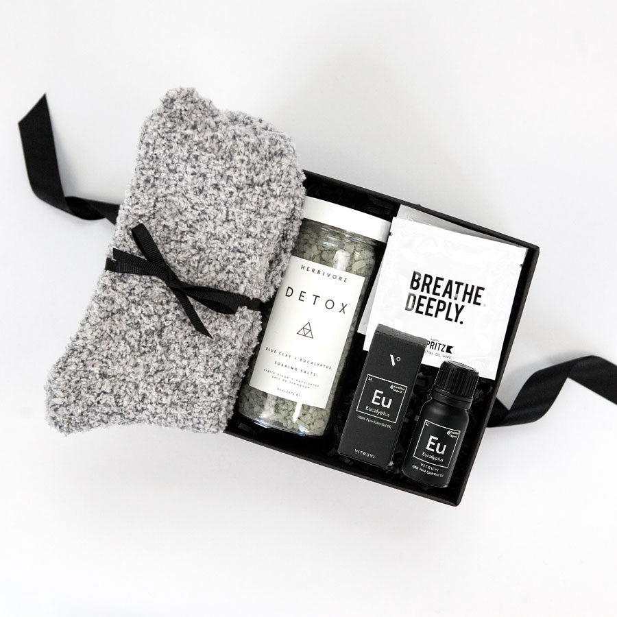 Gray &#39;foxblossom&#39; socks with a black ribbon, &#39;herbivore&#39; detox soaking salts in a glass jar, two &#39;breathe deeply&#39; wipes in a white package, and black eucalyptus essential oil box and bottle, placed in a small black &#39;foxblossom&#39; box with a black ribbon