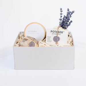 Light grey box containing Potager soap co. lavender soap wrapped in white paper closed with brown twine and purple "PS" wax seal. Lavender bath bomb wrapped in white tissue paper with floral filigree design and purple "PS" wax seal, dried lavender bundle, gold candle tin with white "lavender" sticker.