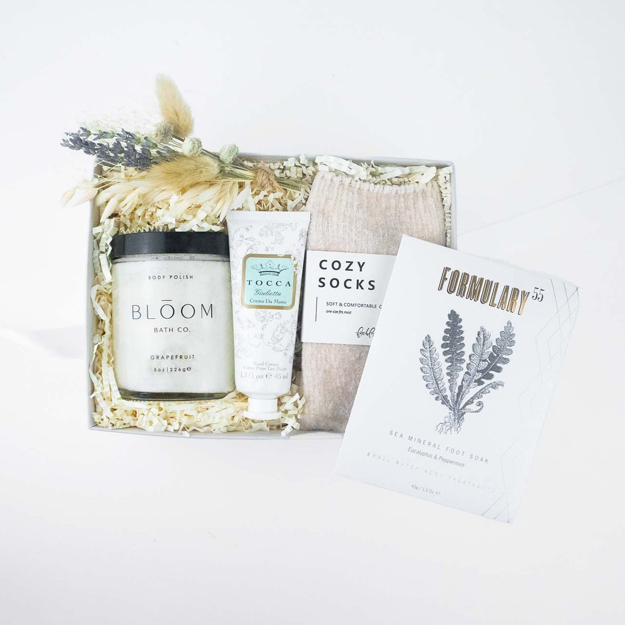 Light grey box containing nude chenille cozy socks,  glass jar of bloom bath co grapefruit body scrub, a dried floral bundle, mini tube of tocca giulietta hand cream with golden floral filigree design, packet of formulary 55 peppermint and eucalyptus sea mineral foot soak.