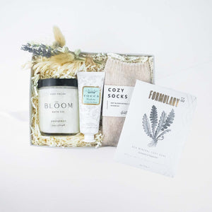 Nude chenille cozy socks,  glass jar of bloom bath co grapefruit body scrub, a dried floral bundle, mini tube of tocca giulietta hand cream with golden floral filigree design, packet of formulary 55 peppermint and eucalyptus sea mineral foot soak arranged in a light grey box with white paper shred.