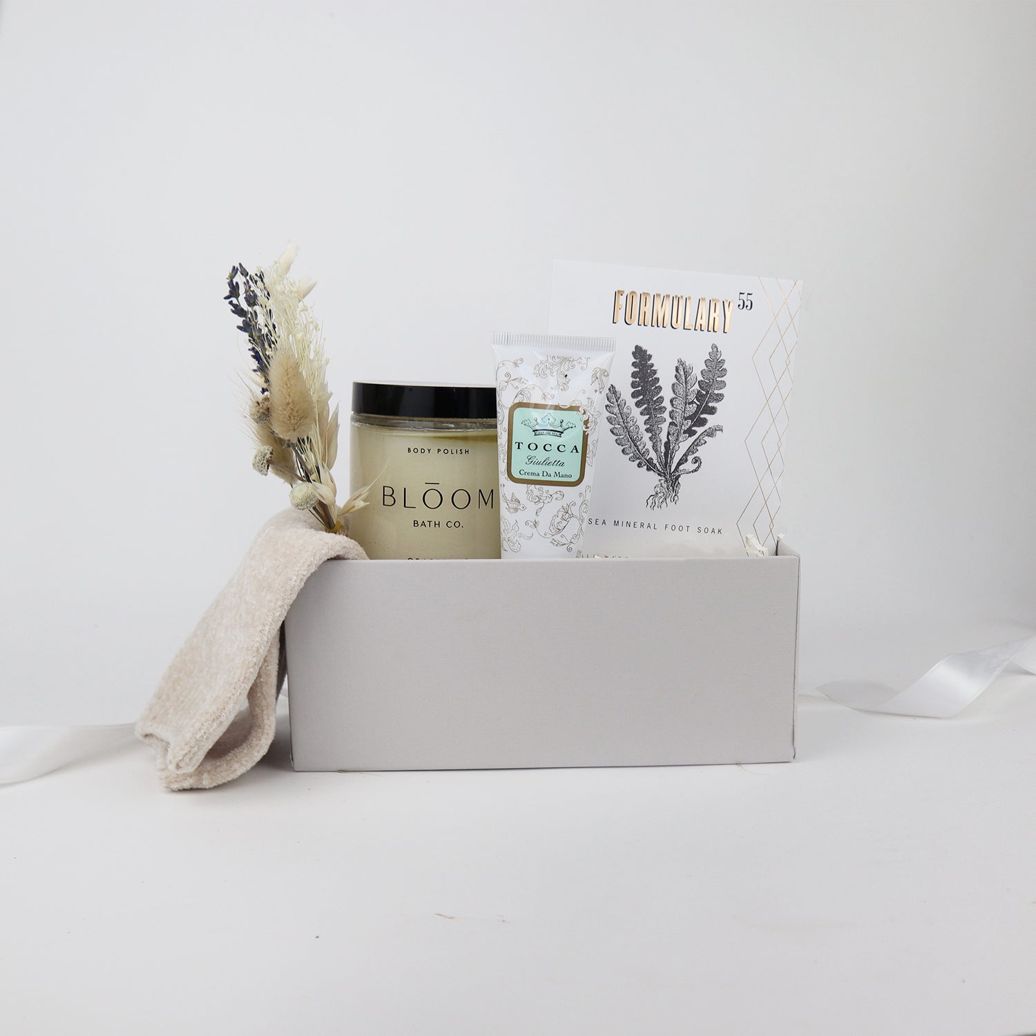 Nude chenille cozy socks,  glass jar of bloom bath co grapefruit body scrub, a dried floral bundle, mini tube of tocca giulietta hand cream with golden floral filigree design, packet of formulary 55 peppermint and eucalyptus sea mineral foot soak arranged in a light grey box with white paper shred.