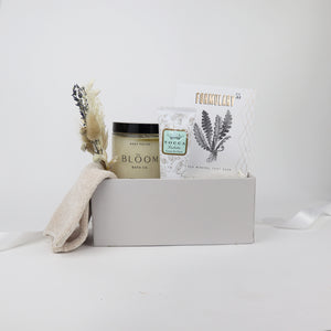 Light grey box containing nude chenille cozy socks,  glass jar of bloom bath co grapefruit body scrub, a dried floral bundle, mini tube of tocca giulietta hand cream with golden floral filigree design, packet of formulary 55 peppermint and eucalyptus sea mineral foot soak.