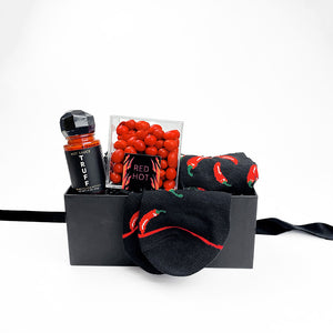 Black box containing a glass bottle of Truff hot sauce with black plastic faceted cap, clear plastic cube containing red hot cinnamon candies, a pair of black socks with red chili peppers design.