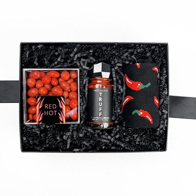 Black box with black paper shred containing a glass bottle of Truff hot sauce with black plastic faceted cap, clear plastic cube containing red hot cinnamon candies, a pair of black socks with red chili peppers design.