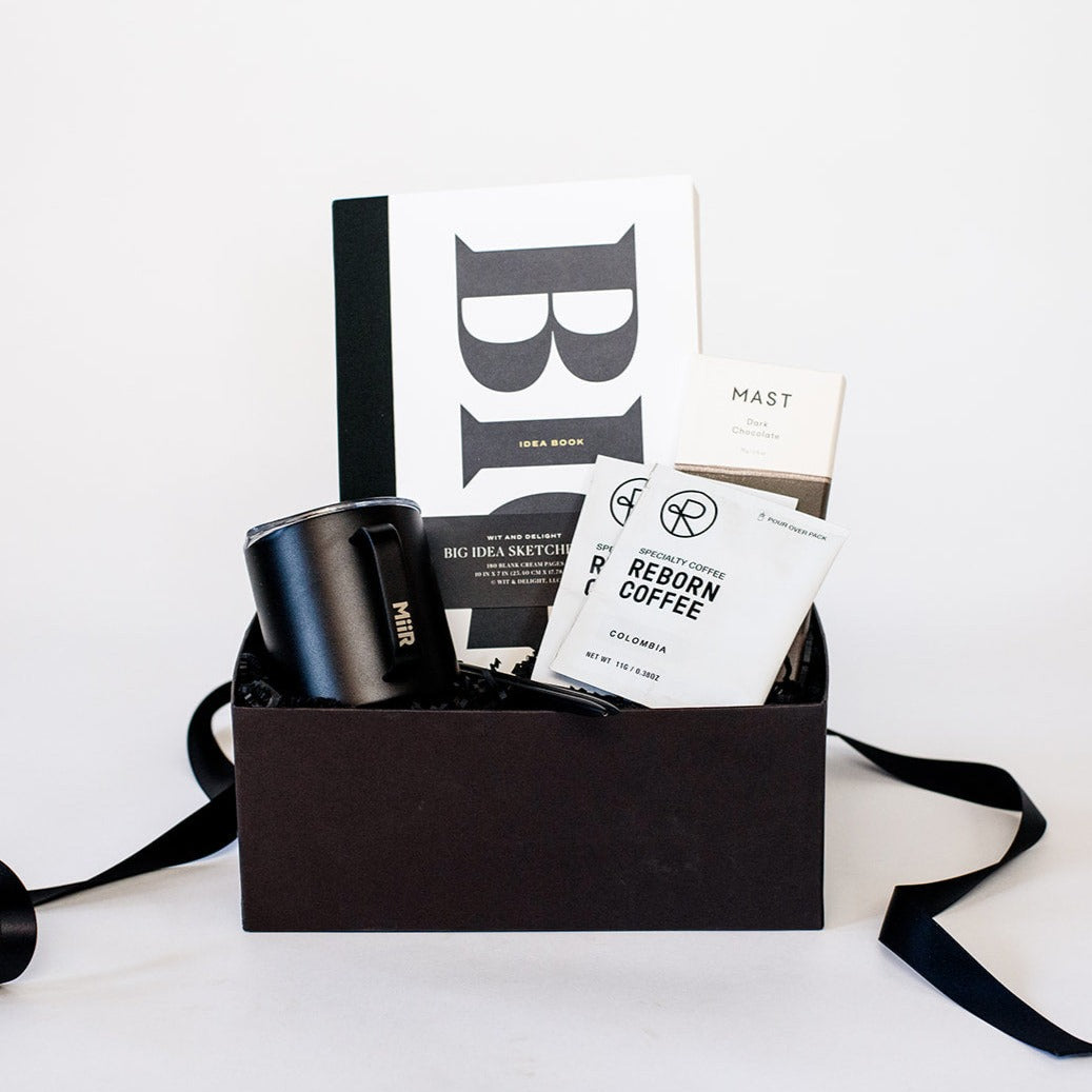 Wit and Delight Big Idea Box, a black Miir camp mug with a lid, two black pens, two packets of Reborn Pour Over Colombia Coffee, and a large Mast dark chocolate bar arranged in a black gifting box with black paper shred.