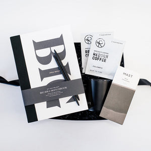 Wit and Delight Big Idea Box, a black Miir camp mug with a lid, two black pens, two packets of Reborn Pour Over Colombia Coffee, and a large Mast dark chocolate bar arranged in a black gifting box with black paper shred.