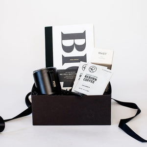 A black box with a Wit and Delight Big Idea Box, a black Miir camp mug with a lid, two black pens, two packets of Reborn Pour Over Colombia Coffee, and a large Mast dark chocolate bar. 